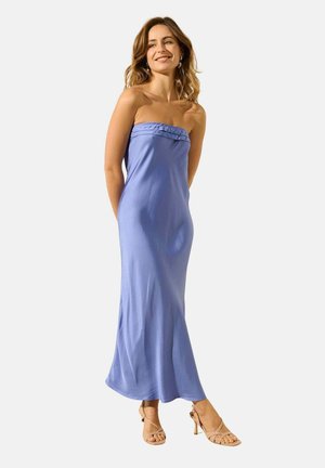 Femme souriante, portant une robe longue bleu clair sans bretelles et des sandales beige à talons hauts, debout les mains derrière le dos.
