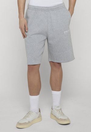 Person iført grå knælange shorts, hvide sneakers og hvide crew-sokker, stående med hænderne i lommerne.