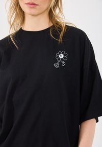 T-shirt de algodão preta com um gráfico branco de uma flor sorridente com pernas no lado esquerdo do peito. Corte descontraído com mangas curtas.
