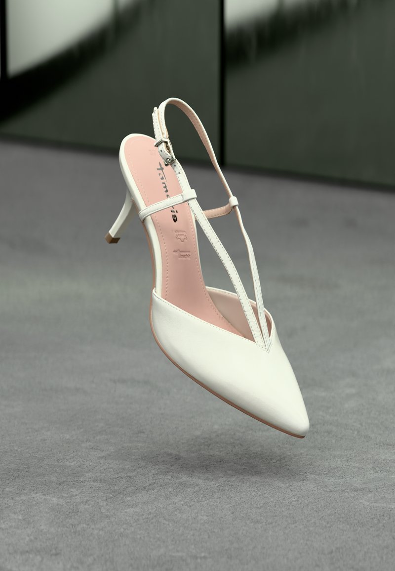 Escarpin blanc à bout pointu avec des brides fines et une semelle intérieure rose, flottant au-dessus d'un sol texturé gris.