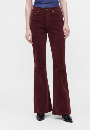 Personne portant un pantalon évasé taille haute rouge foncé, des chaussures noires pointues et un haut bleu rentré, debout devant un fond uni.
