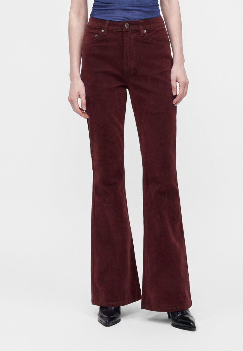 Personne portant un pantalon évasé taille haute rouge foncé, des chaussures noires pointues et un haut bleu rentré, debout devant un fond uni.