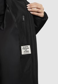 Černý interiér bundy s kapsou na zip a štítkem s nápisem "Teplé objetí vyrobené z látky." Hladký, matný povrch, štíhlý design.