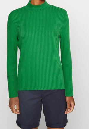 Persoon die een groene geribde trui met lange mouwen en donkerblauwe knielange shorts draagt, tegen een effen achtergrond.