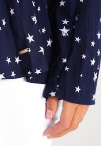 Camicia blu navy con stampa di stelle bianche, con polsini abbottonati e una targhetta metallica con la scritta "Denim". Il tessuto sembra leggero e con una texture.