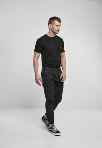 Camiseta negra de manga corta combinada con pantalones cargo negros con bolsillos. Los zapatos son unas zapatillas negras y blancas. El modelo está de perfil.
