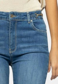 Jeans blu a vita alta in denim con texture liscia, caratterizzati da un bottone frontale, cerniera e hardware decorativo sui lati.