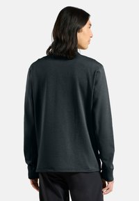 Mann mit schulterlangen, dunklen Haaren, der ein schwarzes Langarm-Shirt und schwarze Hosen trägt, mit dem Rücken zu einer weißen Hintergrundfläche.