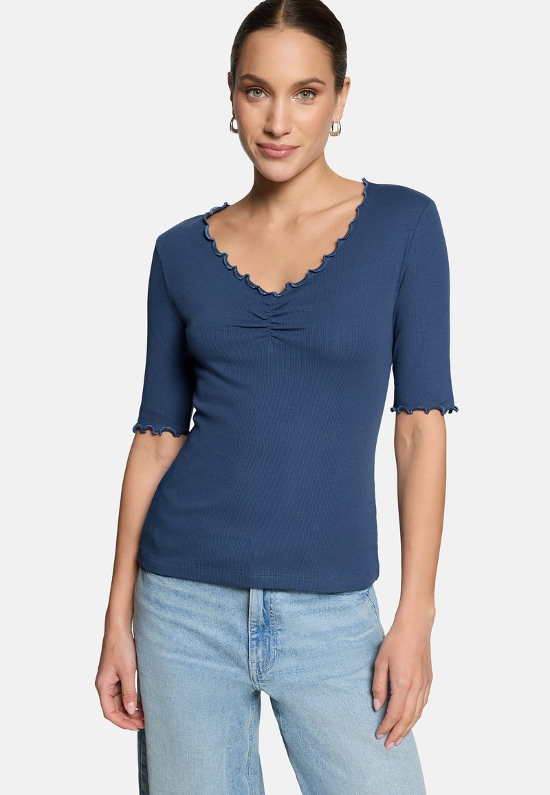 Femme portant un haut bleu marine ajusté avec un col en V et des manches festonnés, associé à un jean bleu clair, posant devant un fond uni.