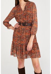 Pitkähihainen oranssi, ruskea ja purppurakuvioinen mekko, jossa on röyhelöhelma ja ruskea vyö, jossa on pyöreä solki.