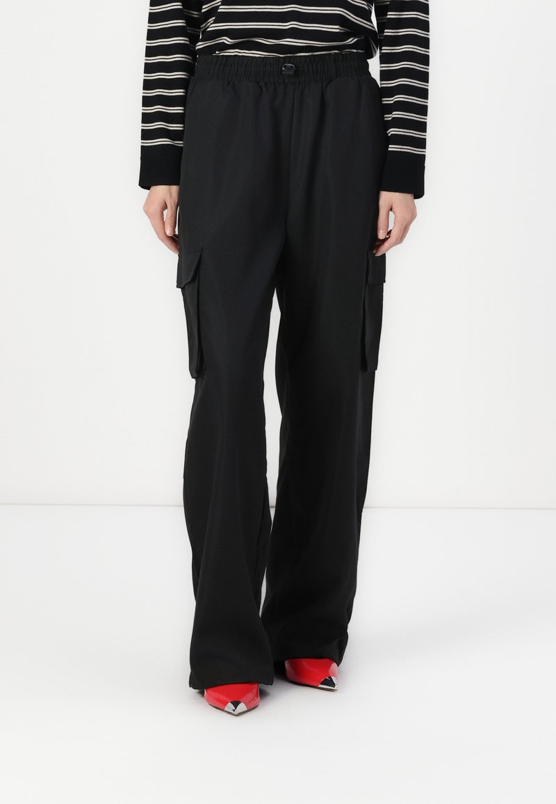 Personne portant un pantalon cargo noir ample avec des poches latérales, un haut à manches longues rayé noir et blanc, et des chaussures rouges pointues.