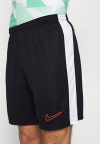 Svarta atletiska shorts tillverkade av lättviktsmaterial, med vita sidopaneler och en orange Nike-logotyp på den nedre framsidan.