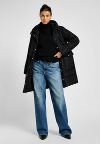 Sort pufferjakke med hætte, der har trykknaplukninger og sidelommer, parret med en sort turtleneck og vide blå jeans.