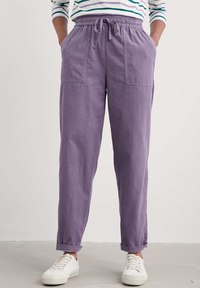Personne portant un pantalon lavande à taille élastique avec poches, revers roulés, baskets blanches et une chemise rayée à manches longues, debout les mains dans les poches.