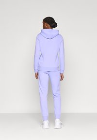 Ανοιχτό μωβ φούτερ με κουκούλα και ταιριαστά παντελόνια φόρμας κατασκευασμένα από απαλό ύφασμα. Το φούτερ διαθέτει κορδόνι και μακριά μανίκια· τα παντελόνια έχουν ελαστικές μανσέτες.