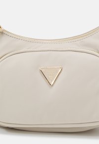 Sac en nylon beige en forme courbe, avec des ferrures dorées et un logo triangulaire. Doté d'une fermeture éclair sur le dessus. Texture lisse.