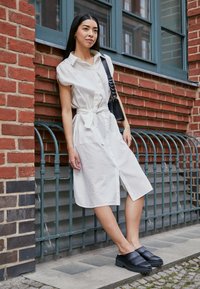 Vestido branco, abotoado, com mangas curtas e detalhe de laço frontal; combinado com sapatos pretos tipo slip-on. O fundo apresenta tijolos e corrimão de metal.