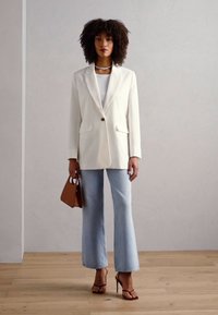 Bílá blazer s jedním knoflíkem, v kombinaci se světle modrými rozšířenými džíny a hnědou kabelkou. Modelka nosí bílý top a sandály na podpatku.