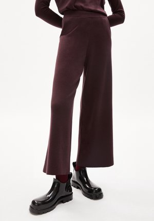 Person trägt dunkelweinrote, weit geschnittene Hosen, passendes Oberteil, weinrote Socken und glänzende schwarze Stiefeletten vor weißem Hintergrund.