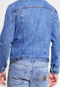 Muž v modré džínové bundě a světle modrých džínách, zobrazený zezadu s rukama volně podél těla.