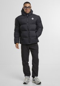 Svart dunjakke med hette, med horisontal quilting, glidelåslukking og sidelommer. Kombinert med svarte bukser og sølvsneakers.