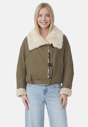 Kvinde iført en olivengrøn jakke med cremefarvet pelskrave og manchetter, kombineret med lyseblå jeans, smilende og kigger til siden.