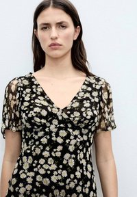 Schwarze florale Bluse mit kurzen, transparenten Ärmeln, die weiße Gänseblümchenmuster aufweist. Der V-Ausschnitt und das leichte Material sorgen für ein sanftes, fließendes Erscheinungsbild.