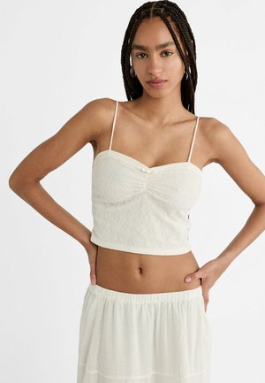 Afgekropte witte kanten top met dunne bandjes, voorzien van een gegatherde voorkant en knoopdetail bij de halslijn. Zachte, textuurrijke stof.