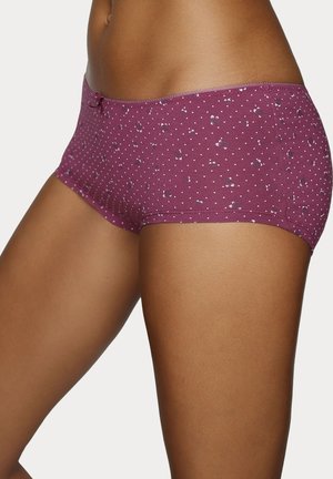 Frau trägt lila Boyshort-Unterwäsche mit kleinen weißen Punkten und dezenten Blumenmustern auf weißem Hintergrund.