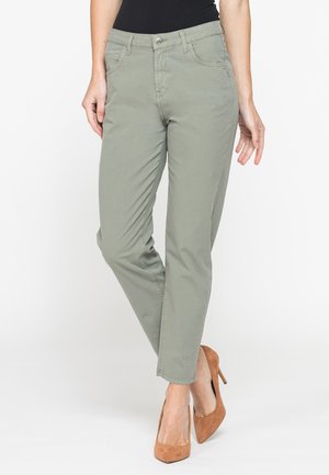 Donna che indossa pantaloni dritti verde chiaro e scarpe marroni con tacco, in piedi con le gambe incrociate su sfondo bianco.