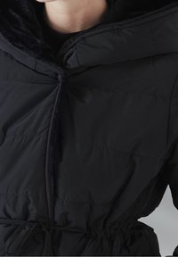 Chaqueta negra acolchada con capucha forrada de piel sintética, cintura ajustable con cordón y cierre de botón. Presenta paneles cosidos y una textura exterior suave.