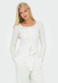 Cardigan blanc à manches longues avec fermeture à boutons, présentant une texture côtelée et un col rond. Accompagné d'un pantalon rayé assorti.