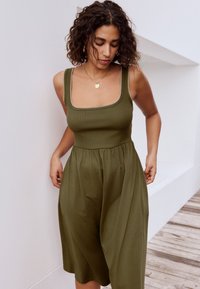 Olivengrøn, ærmeløs kjole med firkantet udskæring, tekstureret stof og samlet talje, med en afslappet pasform, der går til midt på læggen.