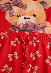 Czerwony zestaw piżamowy z grafiką misia na górze i szortami w kratę z kokardkami. Materiał jest miękki, z elastycznym pasem.