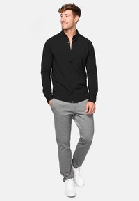 Camisa negra de manga larga con botones y cuello, combinada con pantalones gris claro y zapatillas blancas, de pie con las manos en los bolsillos.