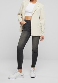 Světle béžový blejzr, bílý crop top, tmavě šedé skinny džíny a bílé tenisky s kovovými akcenty. Džíny mají roztrhané okraje.