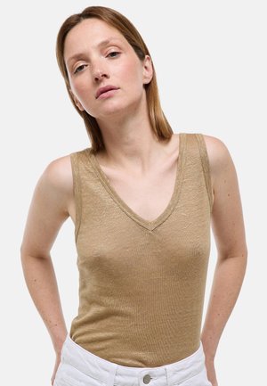Femme aux cheveux châtain clair et raides, portant un haut beige sans manches à col en V et un pantalon blanc, regardant devant elle avec une expression neutre.