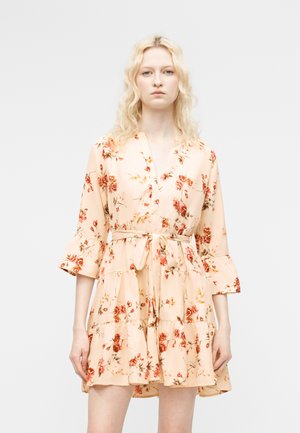 Γυναίκα που φοράει ένα κοντό, μπεζ φόρεμα με λουλούδια, με 3/4 μανίκια που φαρδαίνουν και ζώνη στη μέση δεμένη, στέκεται μπροστά σε ένα απλό φωτεινό φόντο.