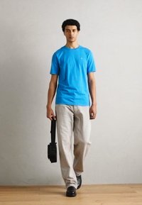 Plavi T-shirt s kratkim rukavima, svjetlosivi traperice, crne cipele, drži crnu torbu. Glatka tekstura tkanine, ležeran dizajn, opušteni kroj.