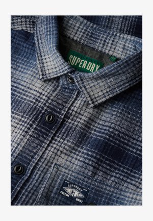 Camicia in flanella a quadri blu e bianca con colletto button-down, bottoni neri e una toppa con logo sulla tasca.