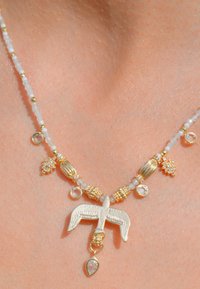 Collier en perles avec des pierres blanches et des accents dorés. Pendentif central en forme d'oiseau avec un détail en forme de goutte en dessous. Design élégant.