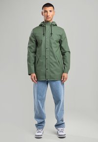 Green waterproof jacket with a hood, snap buttons, and side pockets. Paired with light blue jeans and white sneakers.