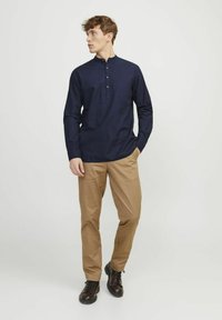 Camicia a maniche lunghe blu navy con mezza pattina e dettagli in bottone, abbinata a pantaloni beige e scarpe nere robuste, in piedi contro uno sfondo chiaro.