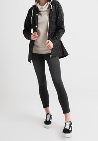 Svart vattentät jacka med subtilt mönster, åtdragen midja och dragsko på huva. Bärs över en beige sweatshirt och matchas med svarta jeans och skor.