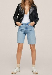 Veste de motard en cuir noir portée sur un débardeur blanc, associée à un short en denim bleu clair. Des baskets blanches avec des accents beige complètent la tenue.