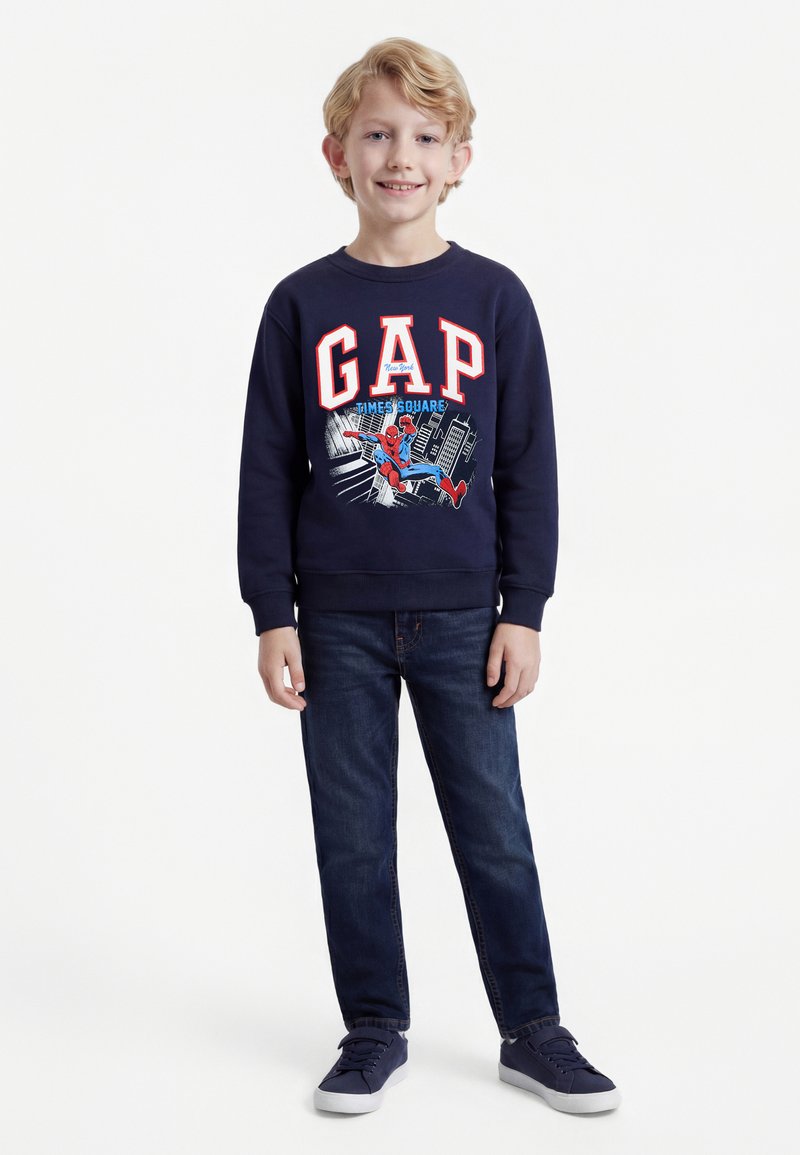 Garçon souriant portant un sweat-shirt bleu marine avec Spider-Man et le logo GAP, un jean foncé et des baskets bleu marine, debout devant un fond blanc.