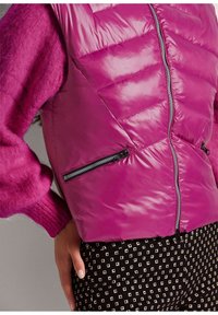 Fuchsia Steppweste mit einem gesteppten Design, Frontreißverschluss und zwei Reißverschlusstaschen. Kombiniert mit gemusterten schwarz-weißen Hosen.