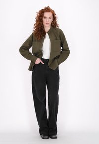 Giacca button-up verde oliva con due tasche sul petto, indossata su una camicia bianca, abbinata a pantaloni neri a vita alta e scarpe nere.