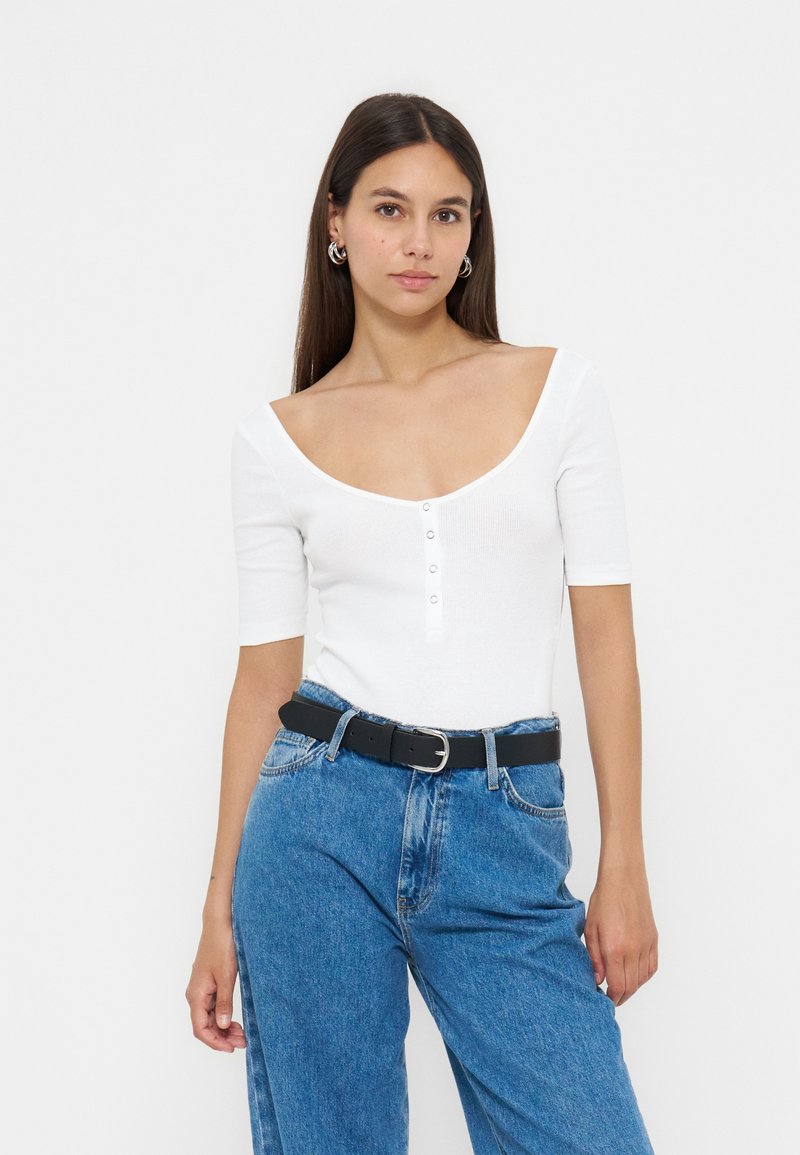 Femme portant un haut blanc à col rond avec des boutons et un jean bleu taille haute, accessoirisée avec des boucles d’oreilles anneaux argentées et une ceinture noire.