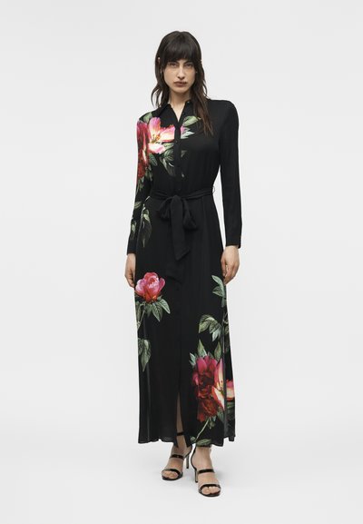 Mujer vistiendo un vestido largo negro con botones, con grandes estampados florales en tonos rosas y rojos, ceñido en la cintura, combinado con tacones negros.